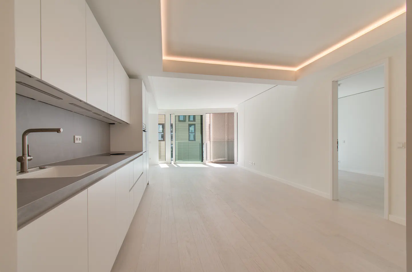 Bright, modern apartment interior with white cabinets, gray countertops, and light wood floors. A balcony door lets in natural light.