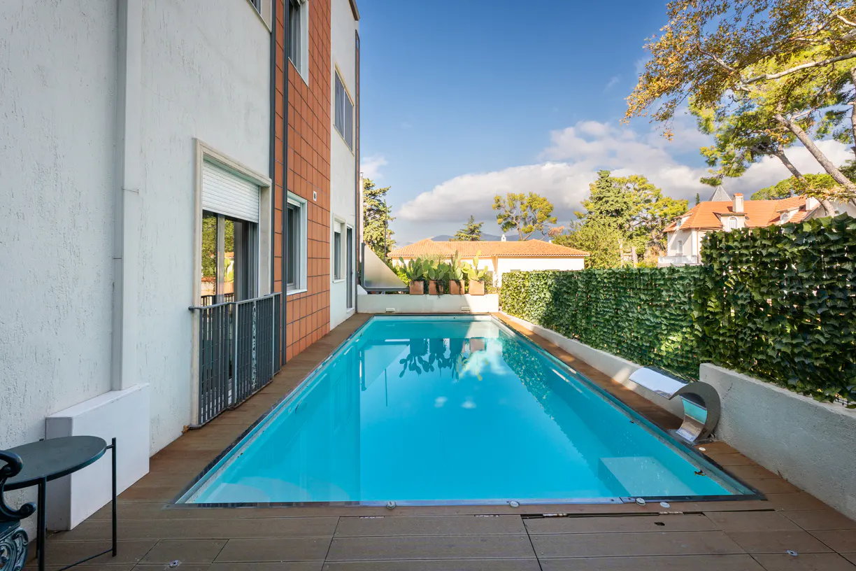 A rectangular, in-ground pool with turquoise water is surrounded by a wooden deck and a white building with brick accents.