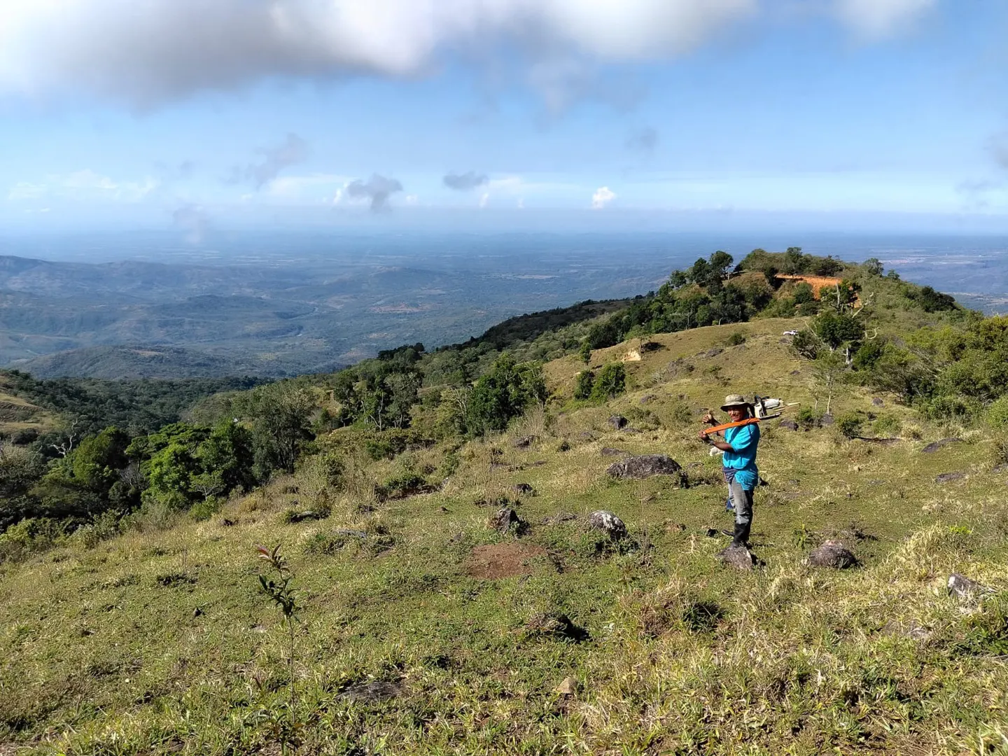 A person in a blue shirt and hat stands on a grassy hillside overlooking a vast, mountainous landscape under a blue sky.