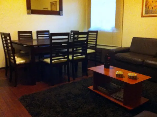 A living and dining area with a dark wood table, chairs, and a brown leather sofa. A red coffee table sits on a dark green rug.