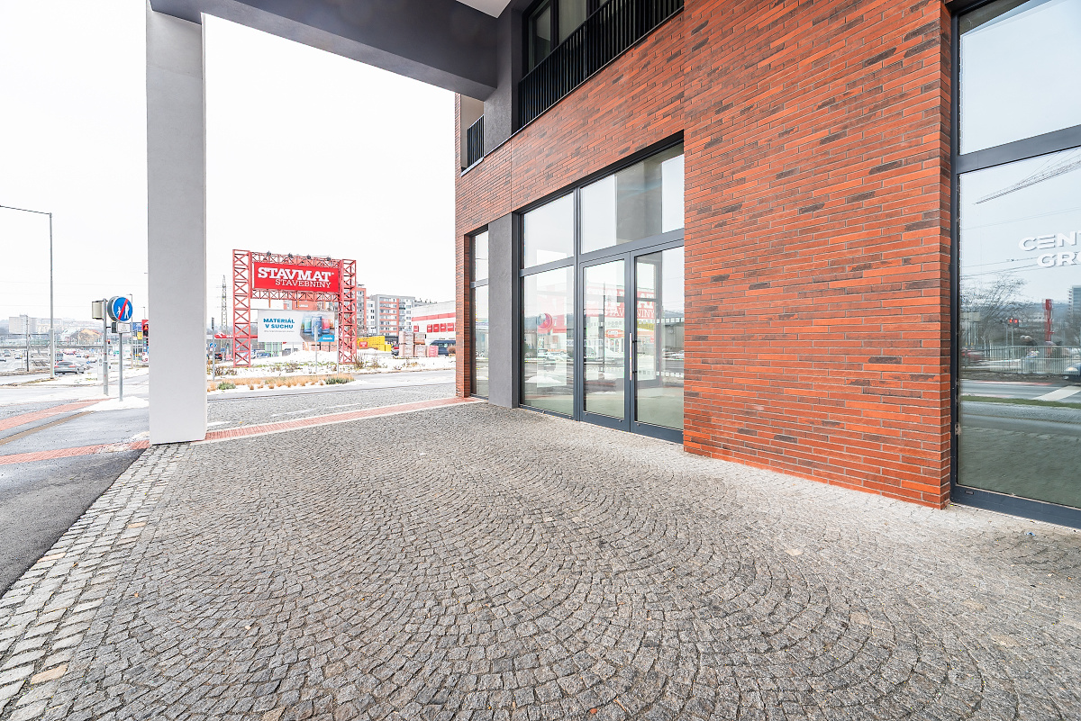 Modern brick building exterior with glass storefronts and a cobblestone entrance, overlooking a street with a red billboard.