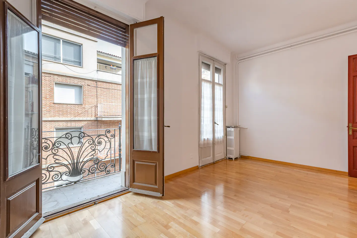 Empty room with light wood floors, white walls, a red door, and open balcony doors overlooking a brick building.