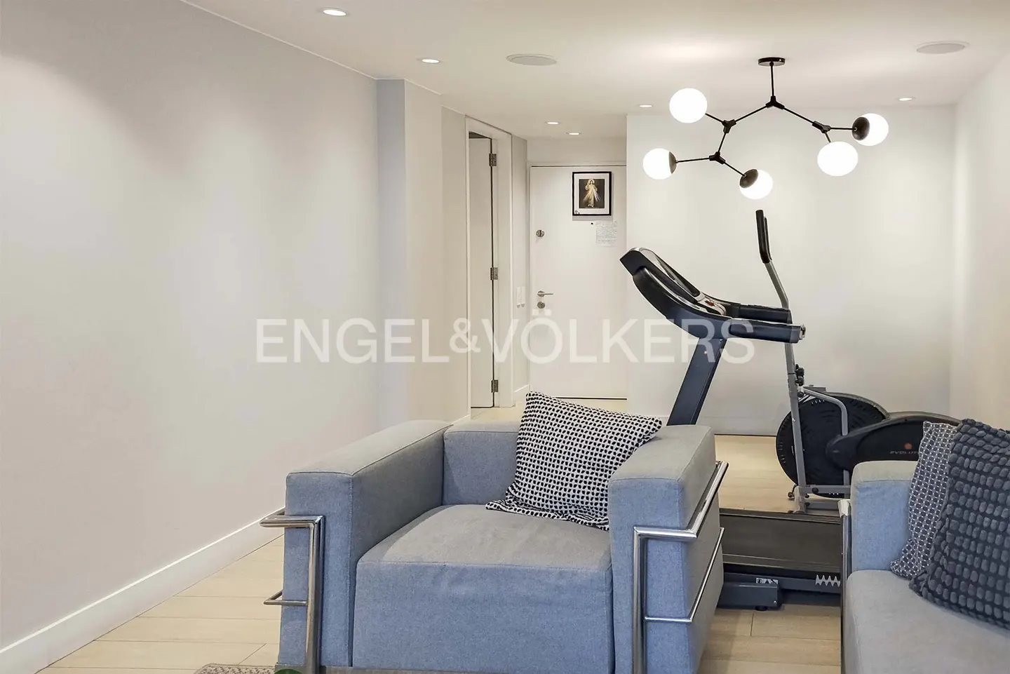 Modern room with a grey armchair, patterned pillows, a treadmill, and a unique black sputnik chandelier.