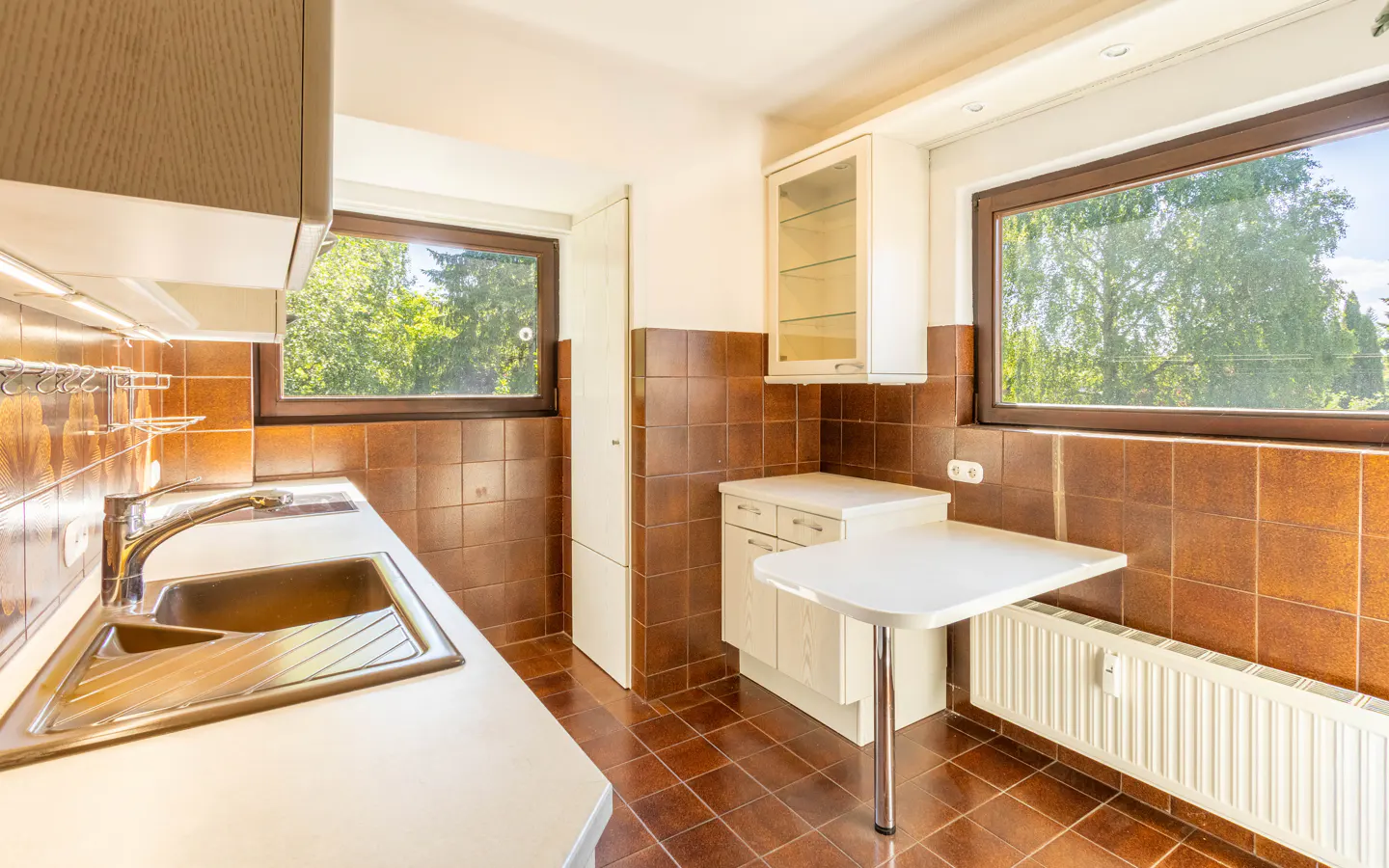 Bright kitchen with brown tiled walls, white cabinets, a stainless steel sink, and a small breakfast table by the window.