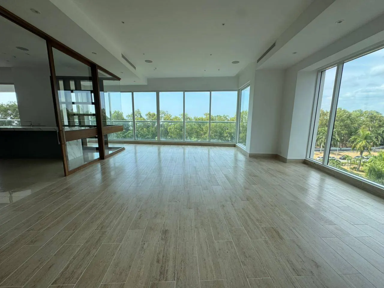 Bright, empty room with light wood-look tile flooring and floor-to-ceiling windows showing trees and sky.