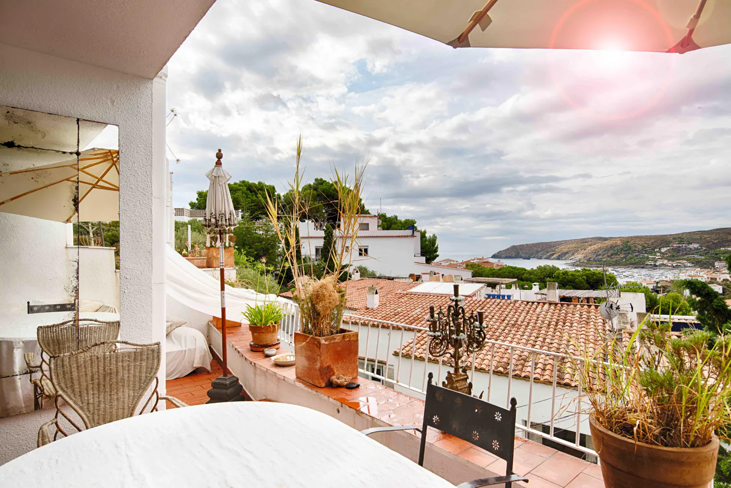 Terrace with wicker chairs, potted plants, and a view of terracotta rooftops overlooking a coastal bay under a cloudy sky.