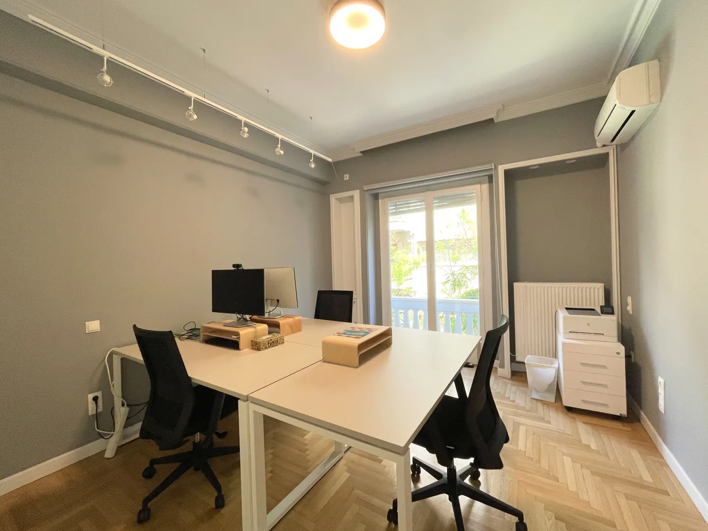 Office space with four desks, black chairs, and computer monitors. A window with a balcony view is in the background.