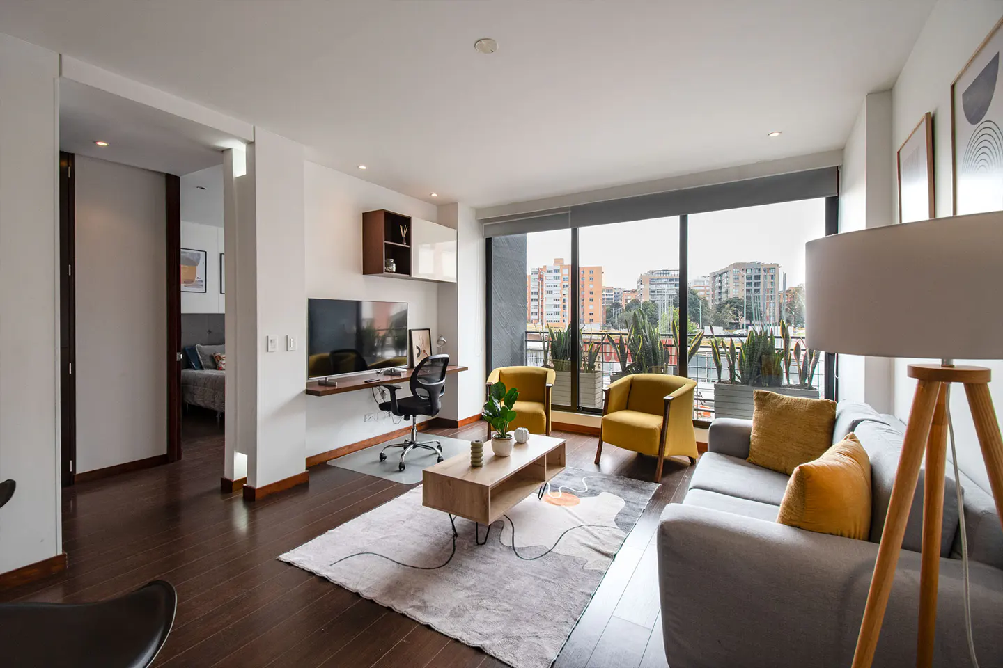 Bright living room with dark wood floors, a gray sofa, yellow chairs, and a desk area by a large window with city views.