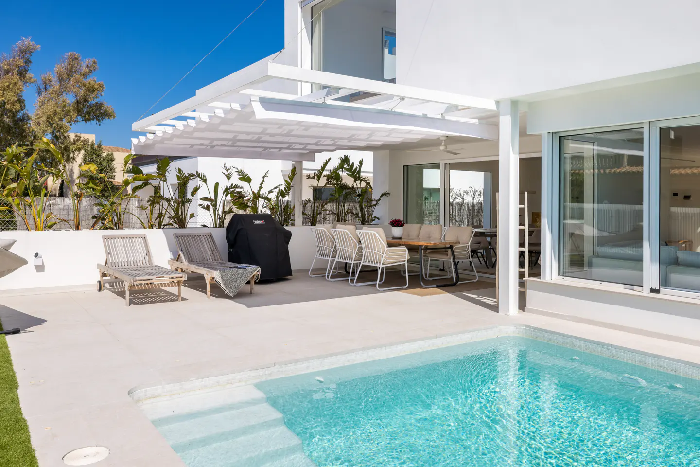 A modern white house with a turquoise pool, lounge chairs, a grill, and an outdoor dining area.