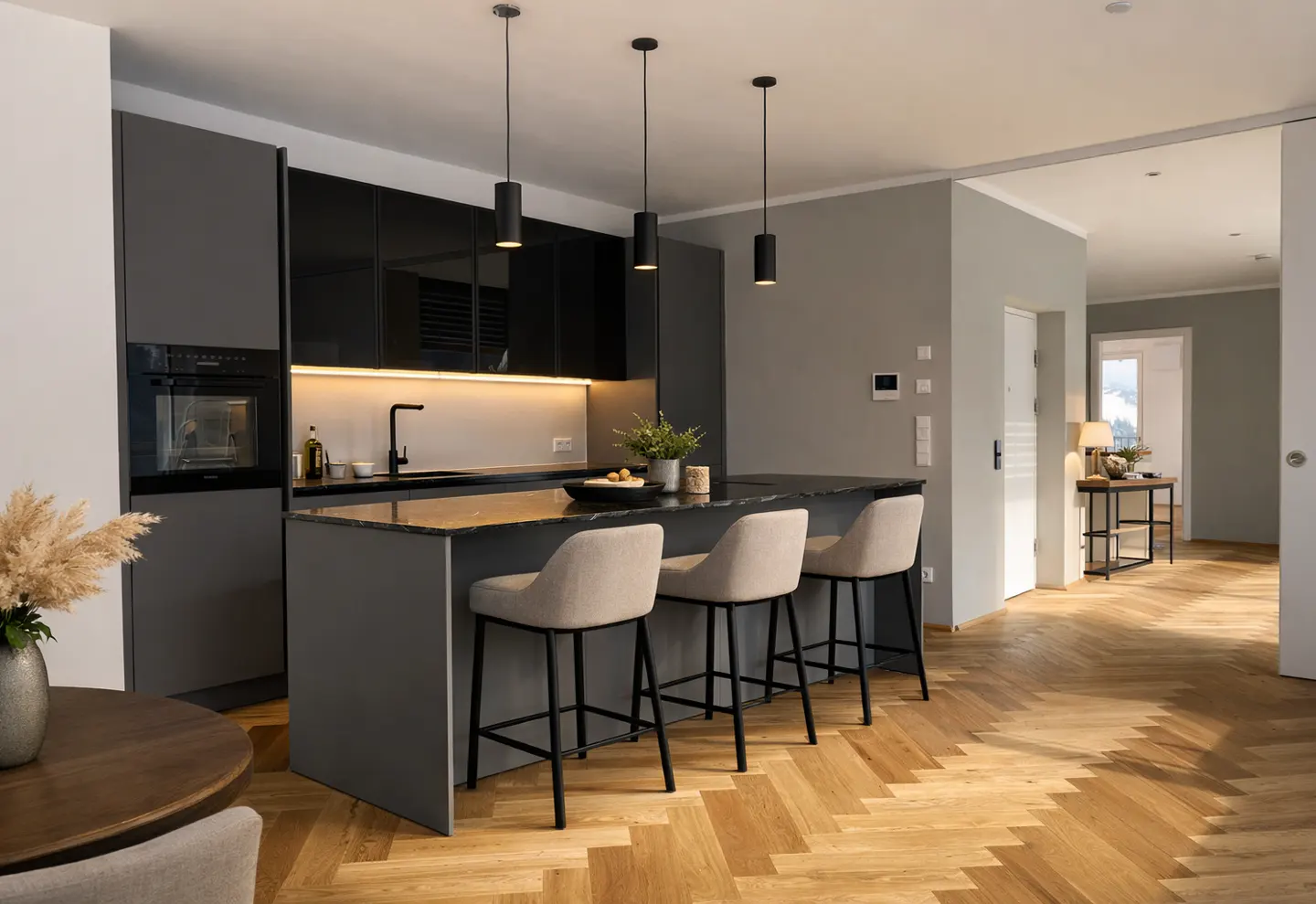 Modern kitchen with gray cabinets, black countertops, and three beige bar stools at the island. Herringbone wood floors and pendant lights.
