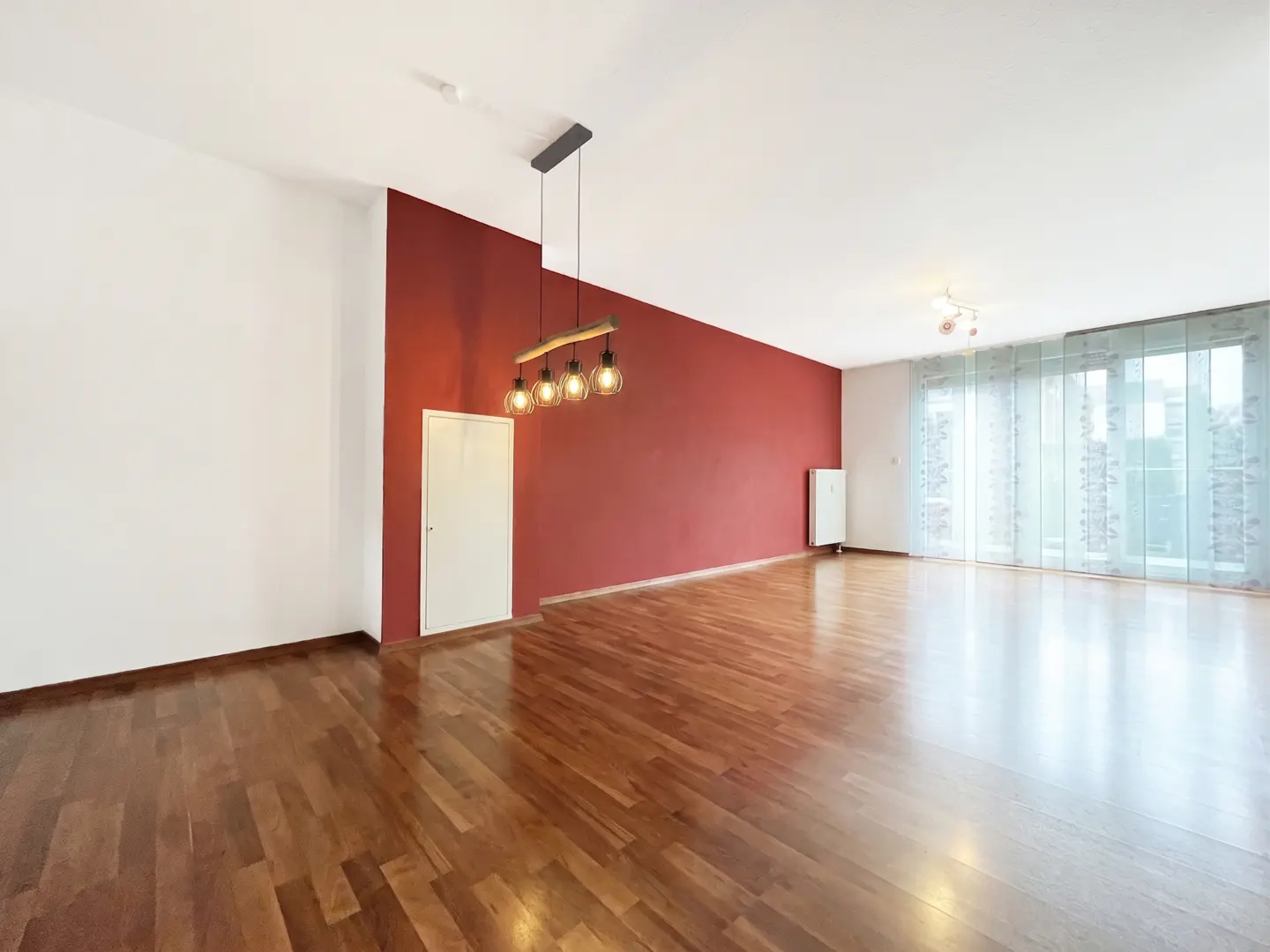 Spacious room with hardwood floors, a bold red accent wall, a modern pendant light, and large windows with sheer curtains.