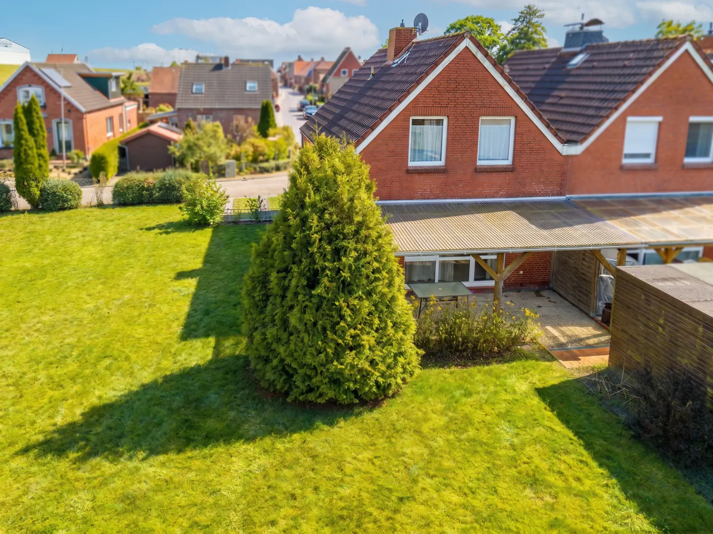 Aerial view of a red brick house with a green lawn, a large evergreen tree, and a covered patio with a table and chairs.