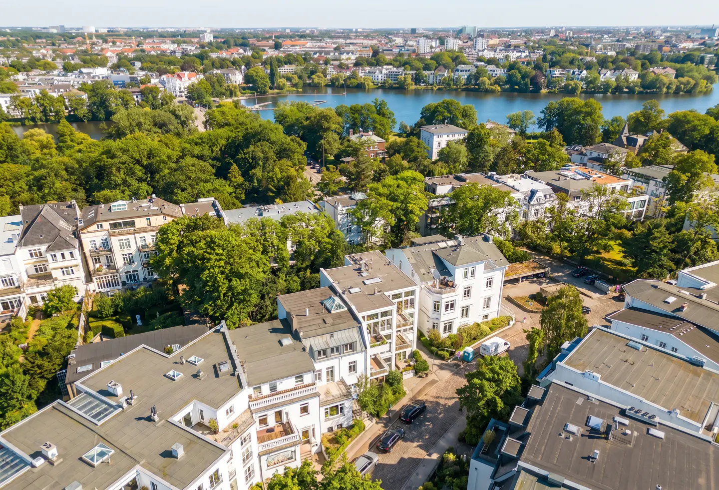 Aerial view of white residential buildings nestled among lush green trees near a lake with a city skyline in the distance.