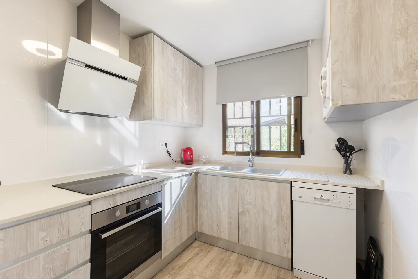 Bright kitchen with light wood cabinets, white countertops, and stainless steel appliances. A window with a gray shade is above the sink.