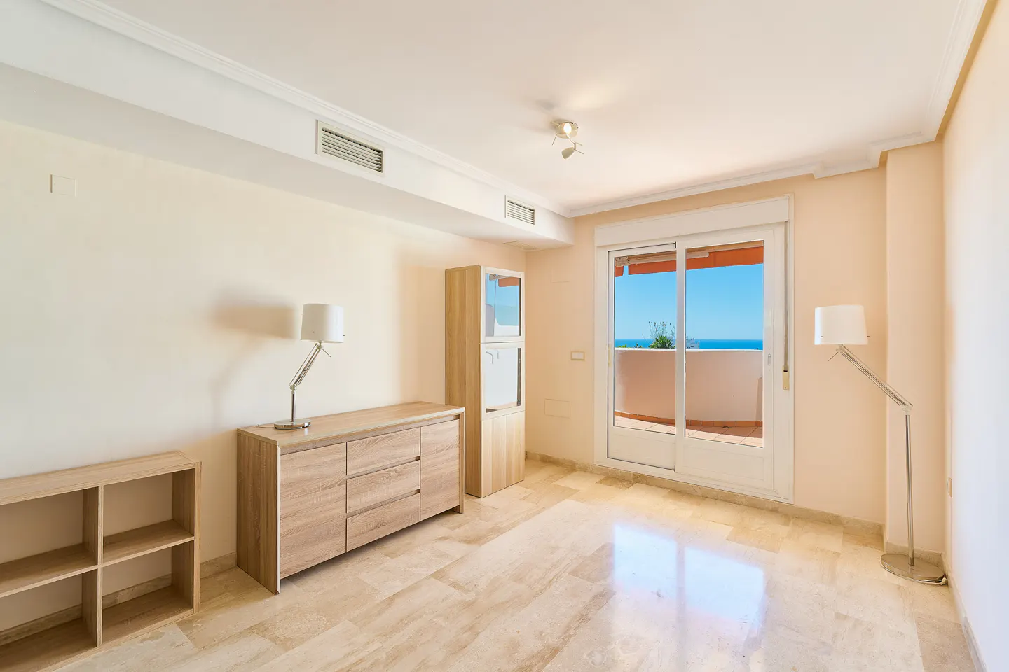 Bright, empty living room with beige walls, marble floors, and light wood furniture. Sliding glass doors lead to a balcony with ocean views.