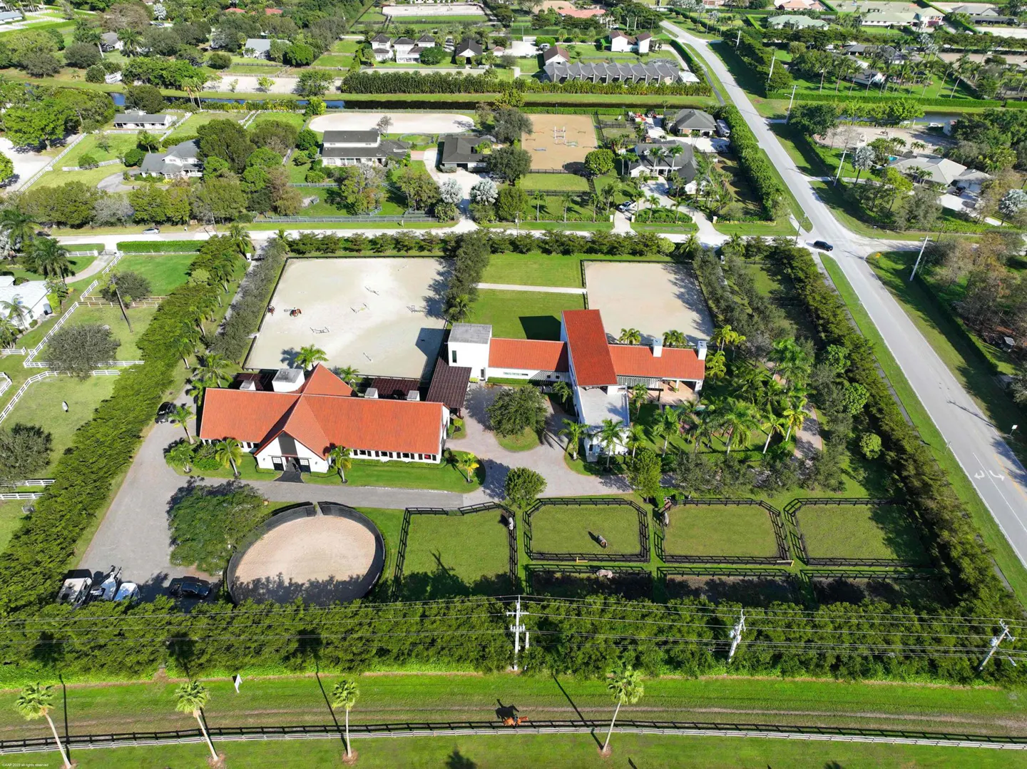 Aerial view of an equestrian estate with red-roofed buildings, horse paddocks, and riding arenas surrounded by green landscaping.