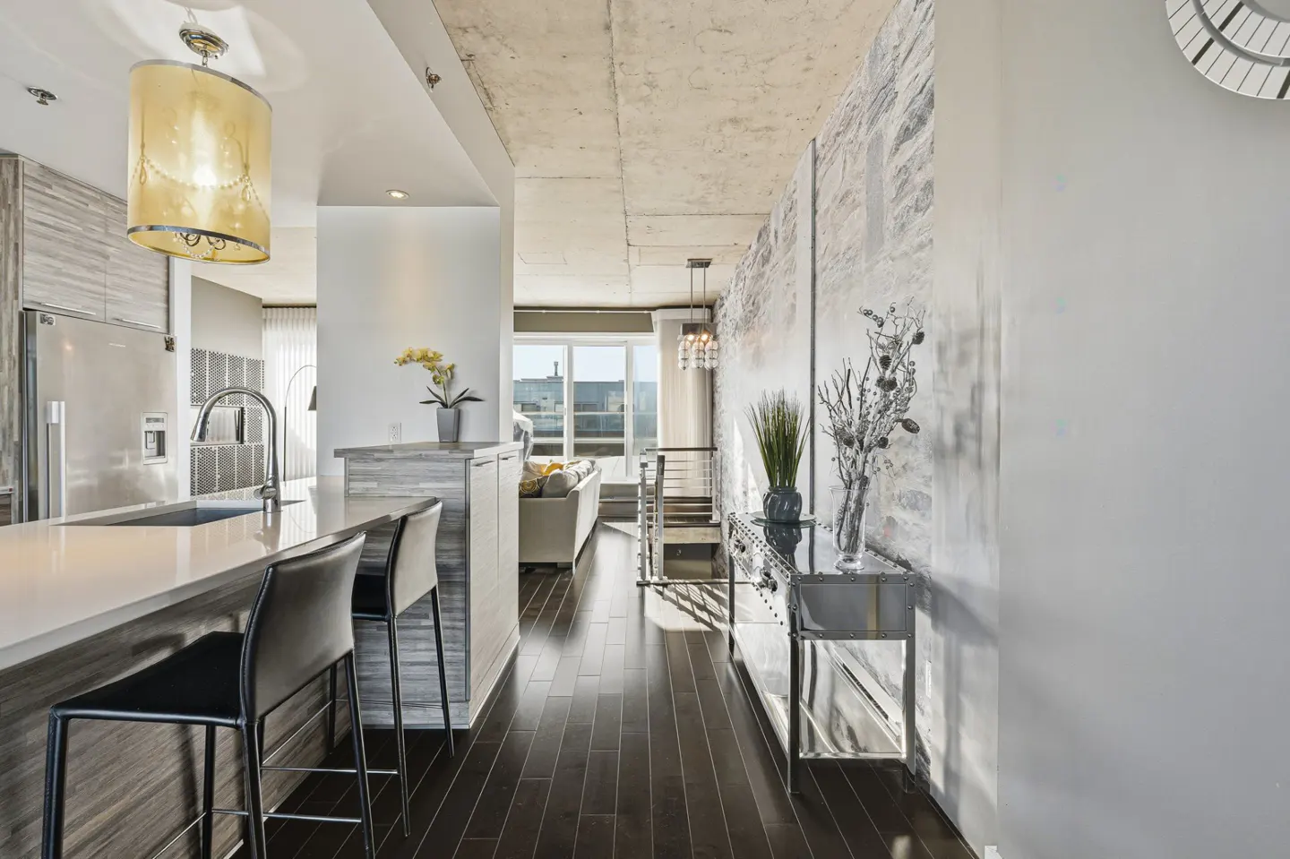 Interior view of a modern apartment with dark wood floors, a kitchen island with black stools, and a living area with a sofa and city view.