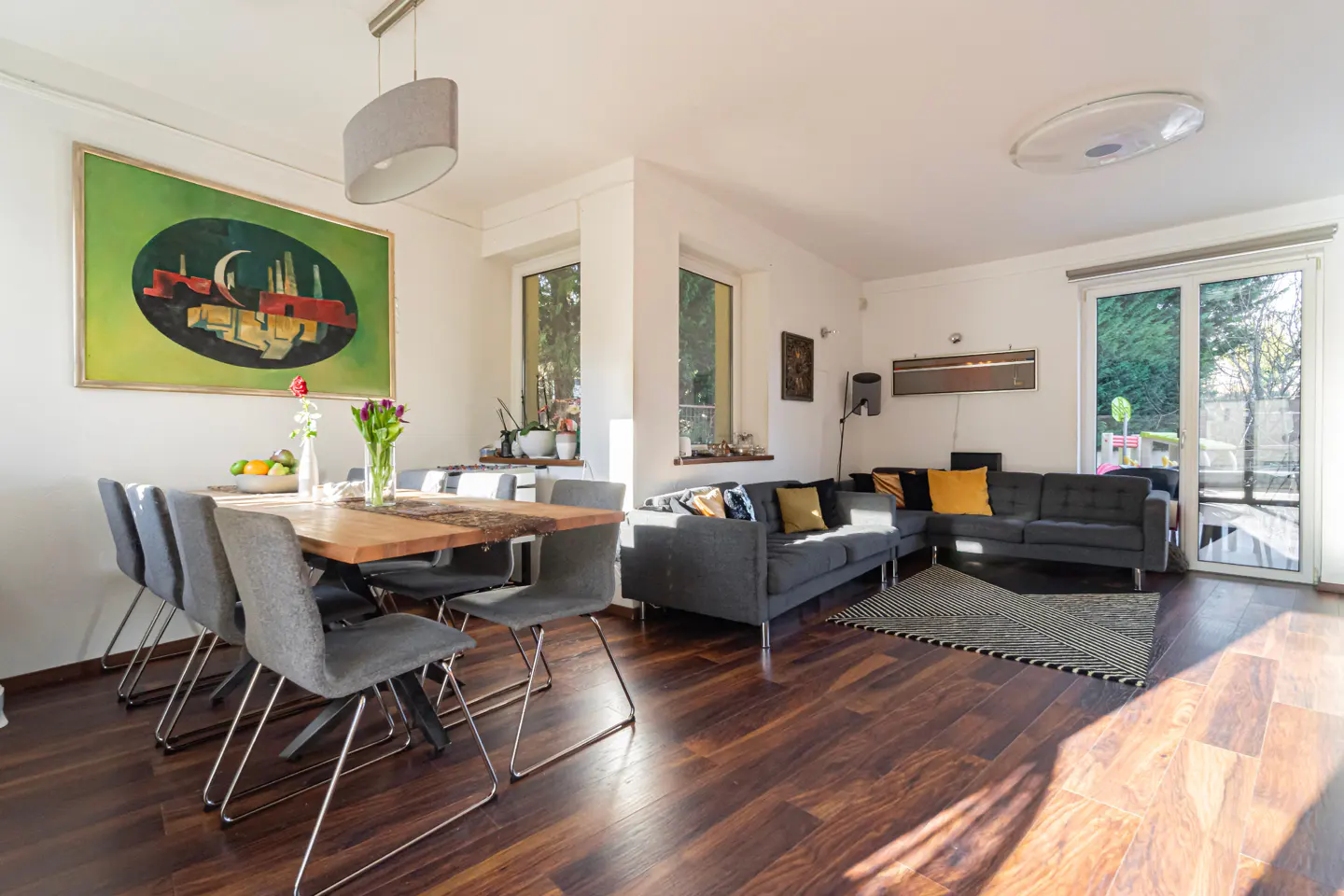 Bright, open-concept living and dining area with wood floors, gray sofas, and a wooden dining table with gray chairs. A large, colorful painting hangs on the wall.