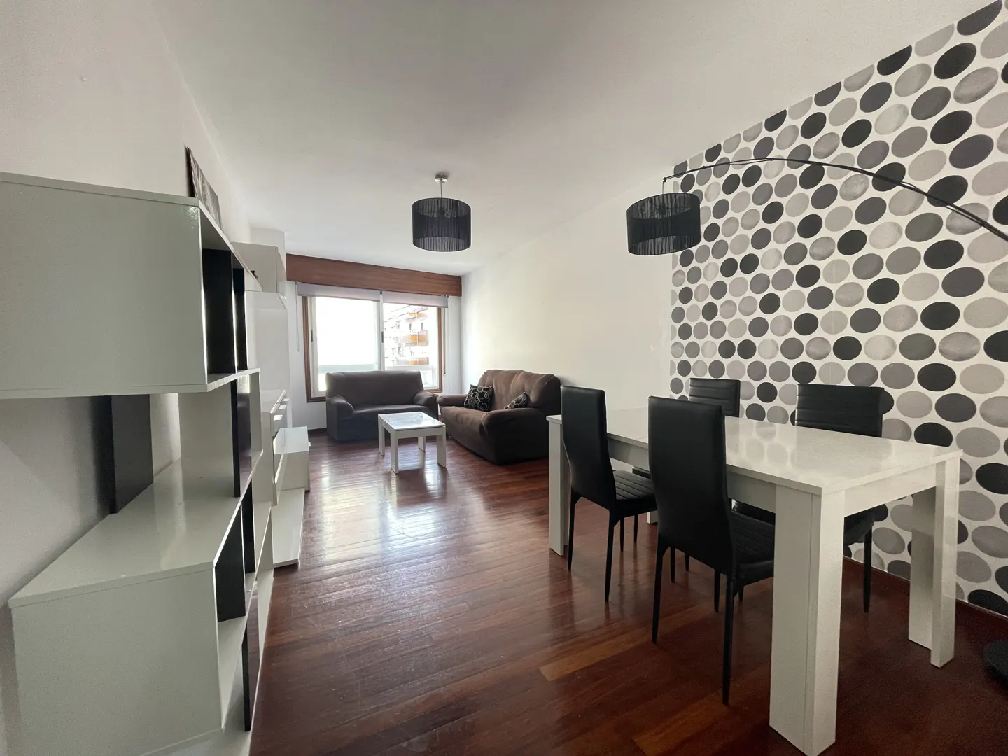 A living room with a white table and black chairs, brown sofas, and a white bookshelf. The wall has a black and white circle pattern.