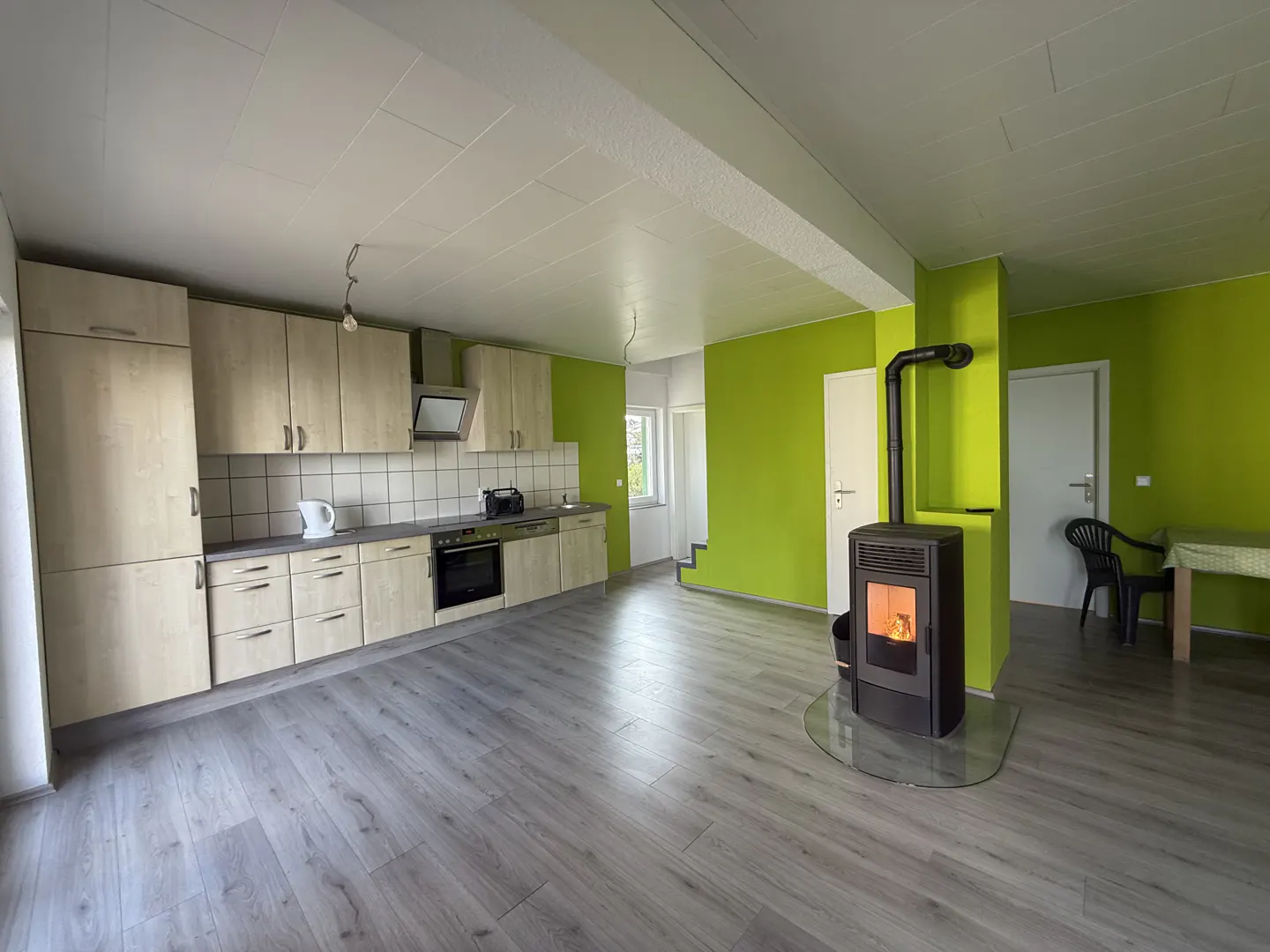 Bright, open kitchen with wood cabinets, gray floors, and green walls. A wood-burning stove sits near a dining area with a black chair.