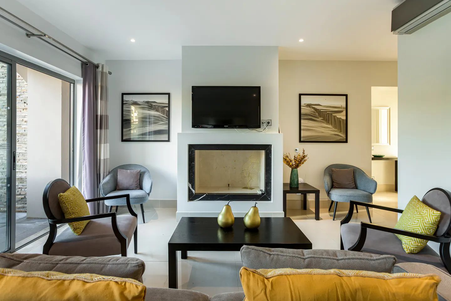 Modern living room with a fireplace, TV, four grey armchairs, a black coffee table with gold pears, and framed wall art.