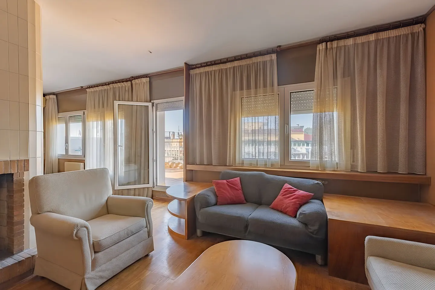 Living room with a gray sofa, red pillows, and a cream armchair. Windows with curtains overlook a city view.