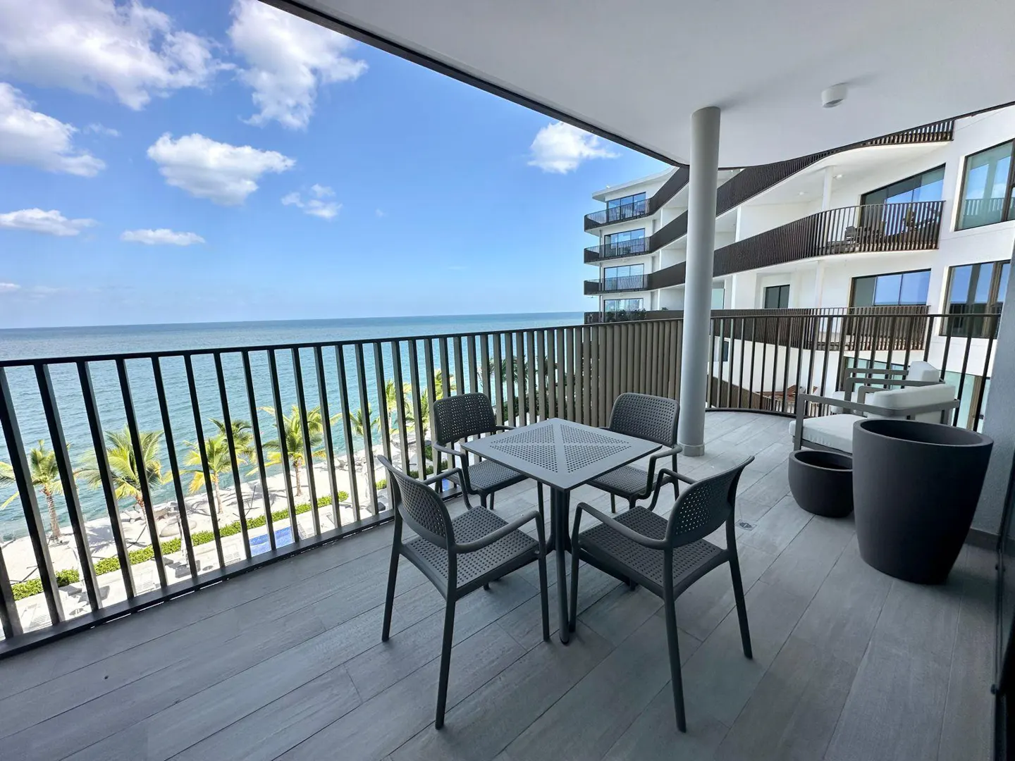 Balcony view with ocean. A gray table and four chairs sit on a gray wood balcony overlooking the ocean and palm trees. A modern building is visible.