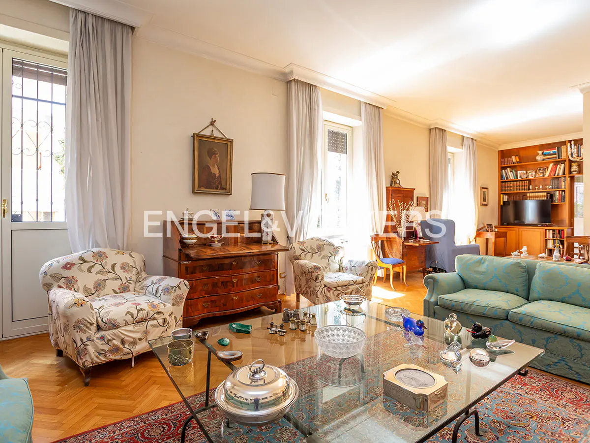 Bright living room with floral armchairs, a blue sofa, and a glass coffee table on a patterned rug. A wooden dresser and framed portrait add character.