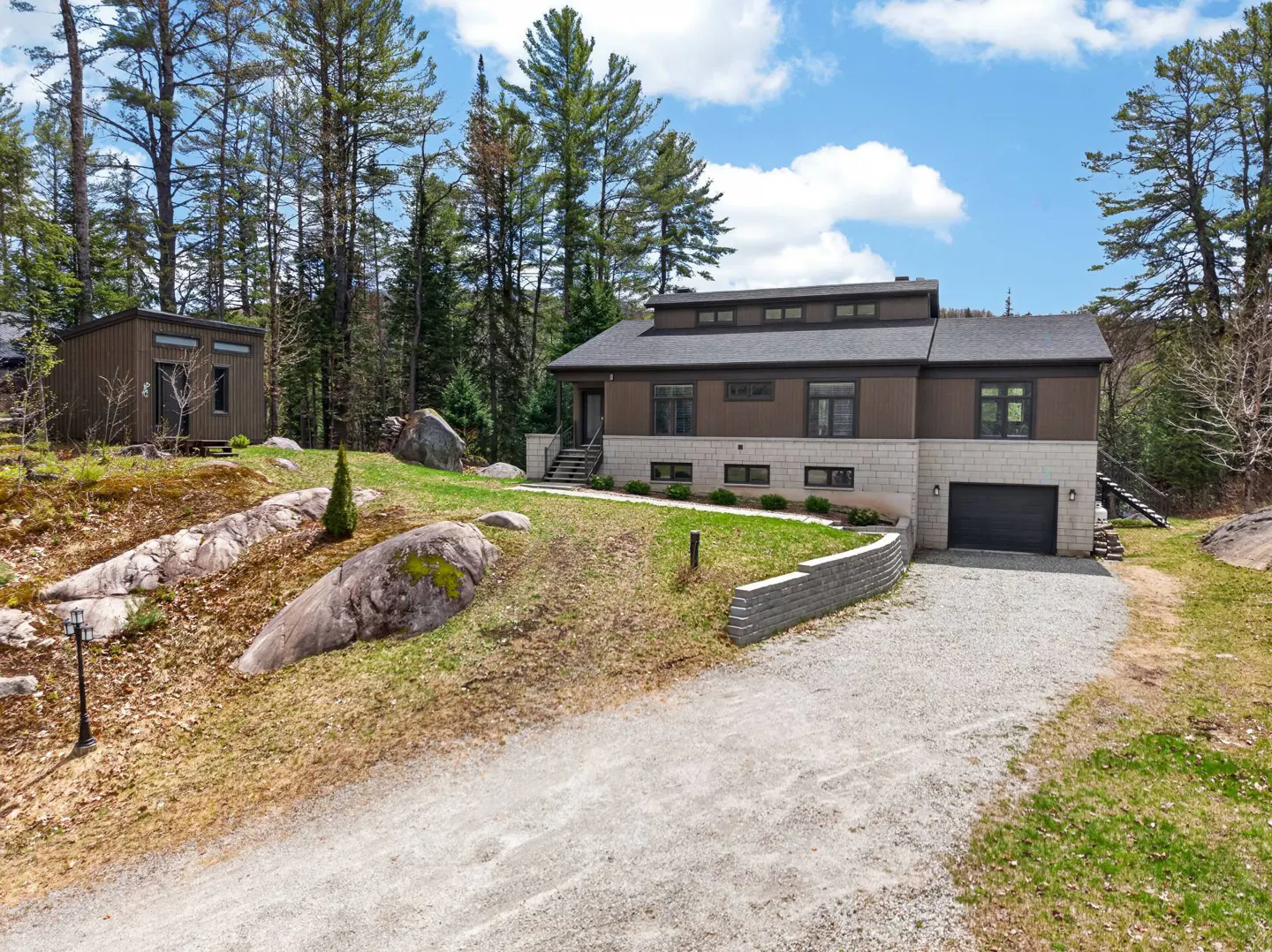 Modern brown house with stone base, black garage door, and gravel driveway, surrounded by trees and rocks.