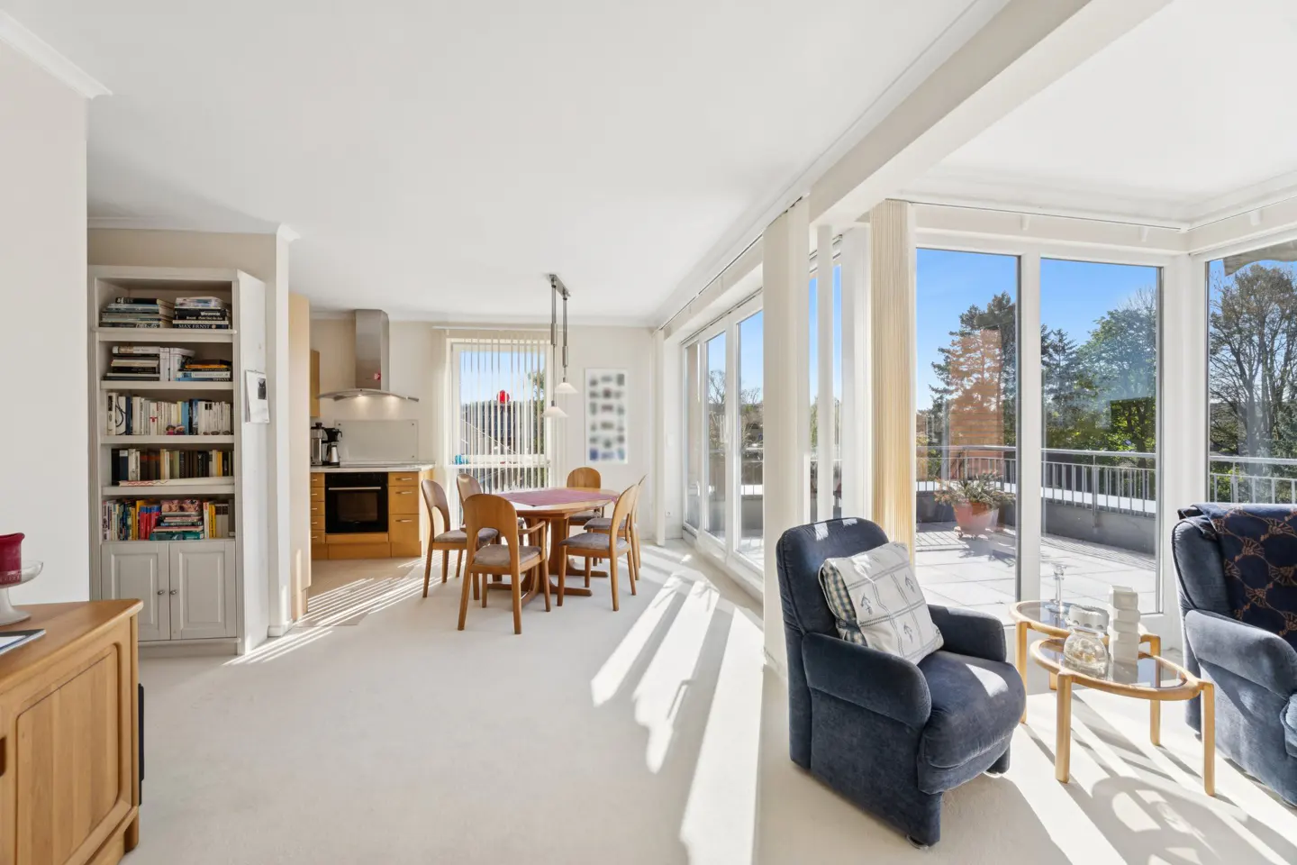 Bright, open-concept living space with a dining table, blue armchairs, and floor-to-ceiling windows overlooking a balcony and trees.