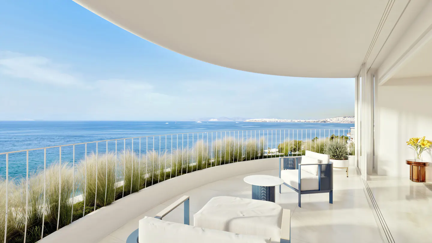 A bright, modern balcony with white furniture overlooks a blue ocean and distant city skyline.