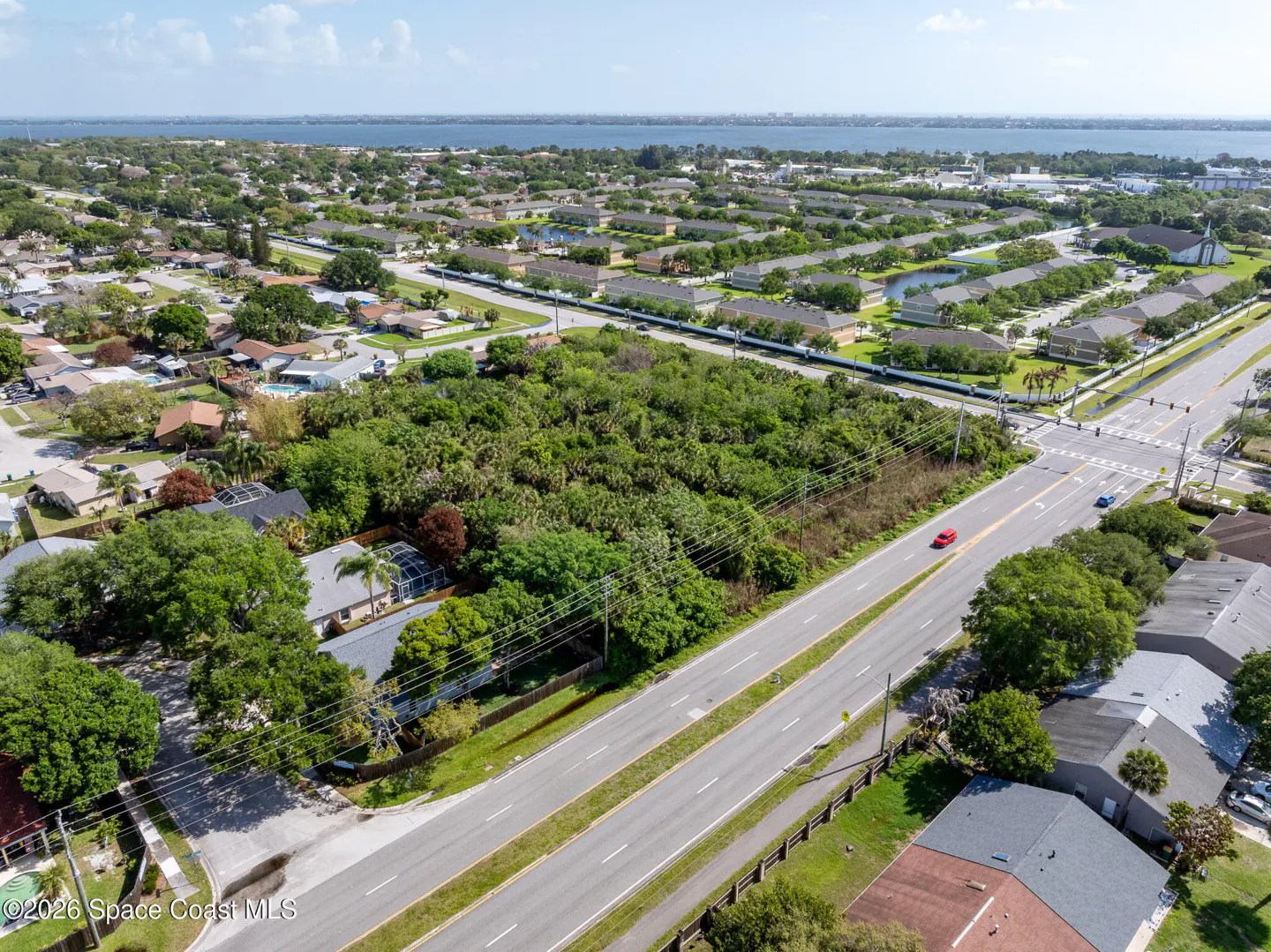 Aerial view of a vacant lot with lush green trees, bordered by a road and residential area under a blue sky.