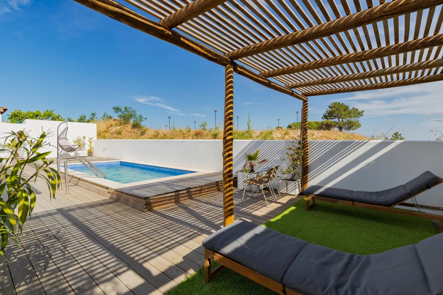 Outdoor patio with pool, lounge chairs, and dining set under a wooden pergola on a sunny day.