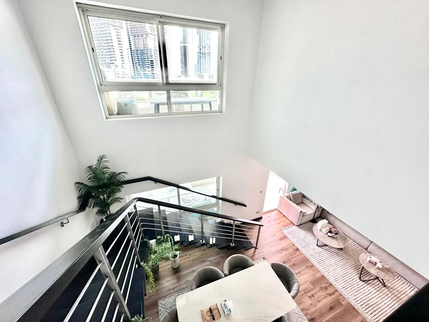 A high-angle shot of a modern, open-concept living space with white walls, wood floors, and a staircase with metal railings. A dining table and living room are visible.