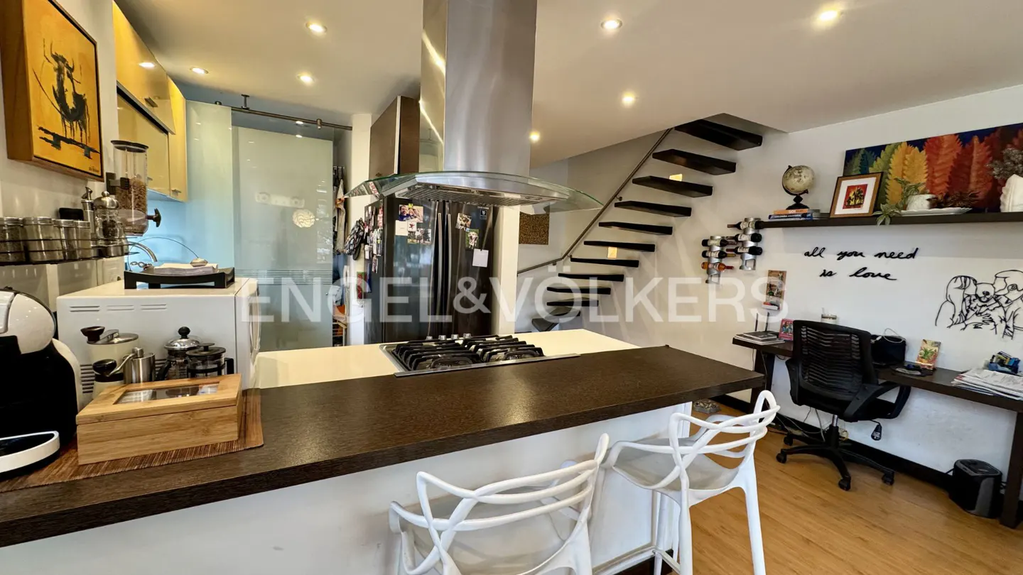 A modern kitchen with a dark countertop island and white chairs. Stainless steel appliances and a floating staircase are visible.
