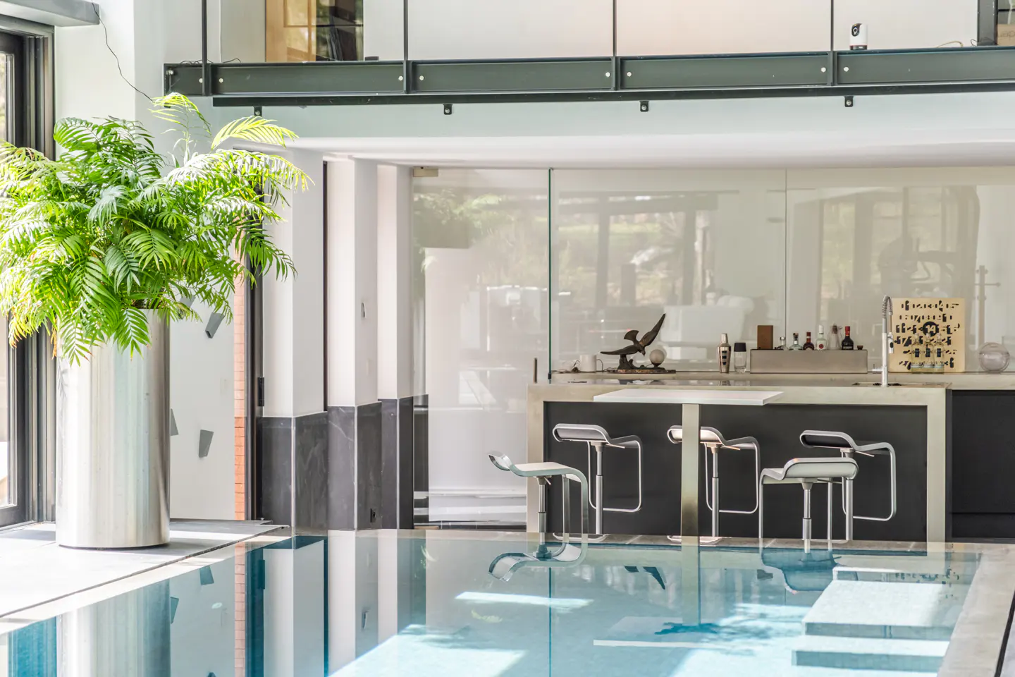 Indoor pool with a bar and stools. A tall plant sits in a silver pot. The water reflects the bar and stools.