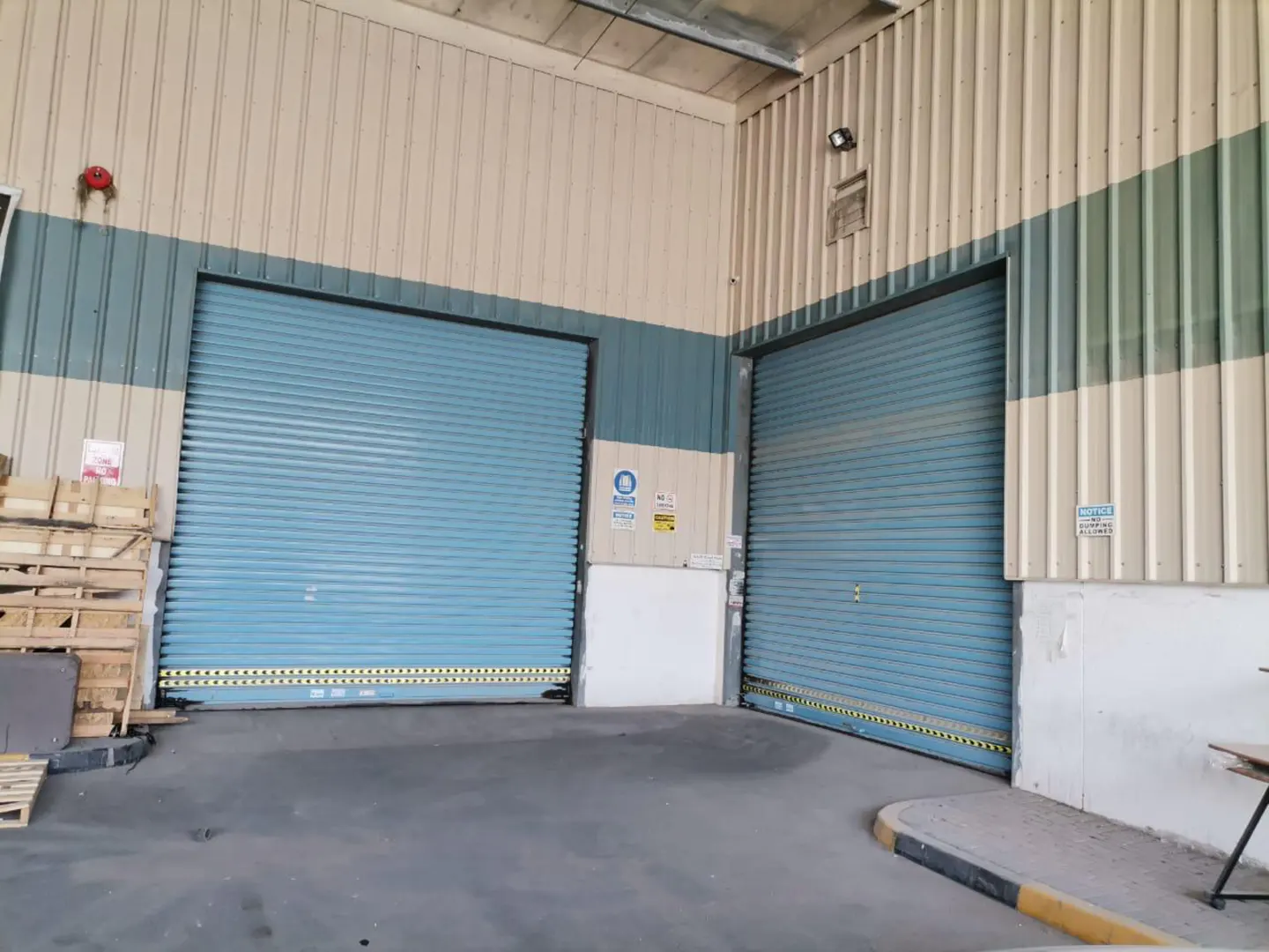 Exterior view of a warehouse with two closed, light blue roll-up doors and a beige and blue/green striped facade.