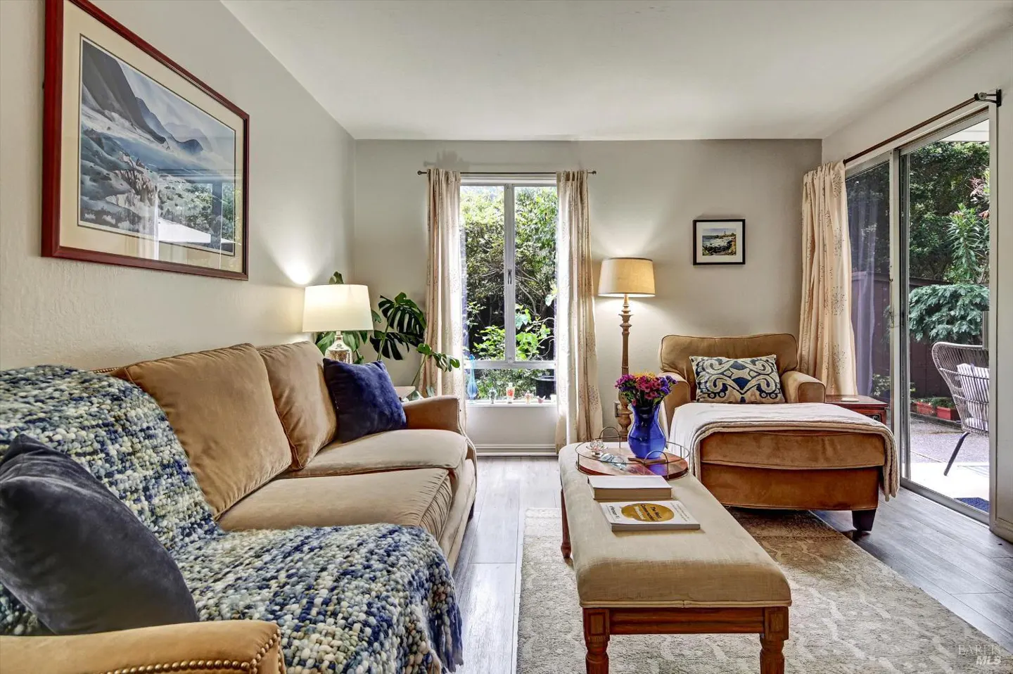 Living room with tan sofa, chaise lounge, and ottoman. A blue throw and pillows add color. A window and sliding door offer views of greenery.