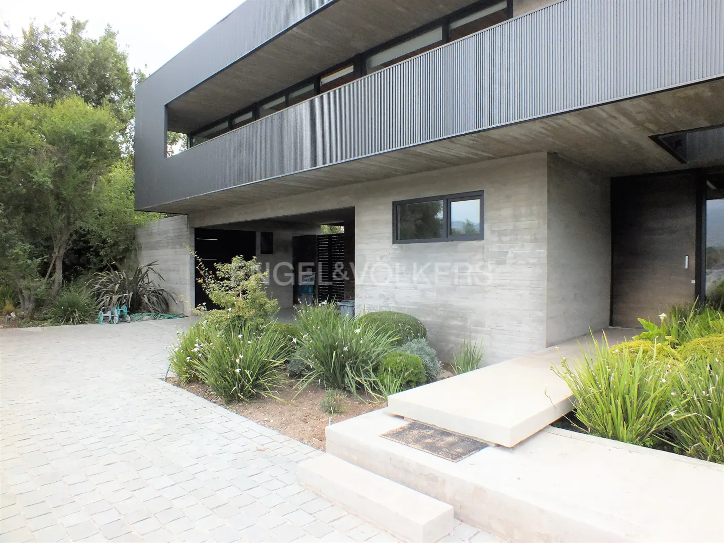Modern two-story home with concrete and gray siding, a paved driveway, and lush green landscaping.