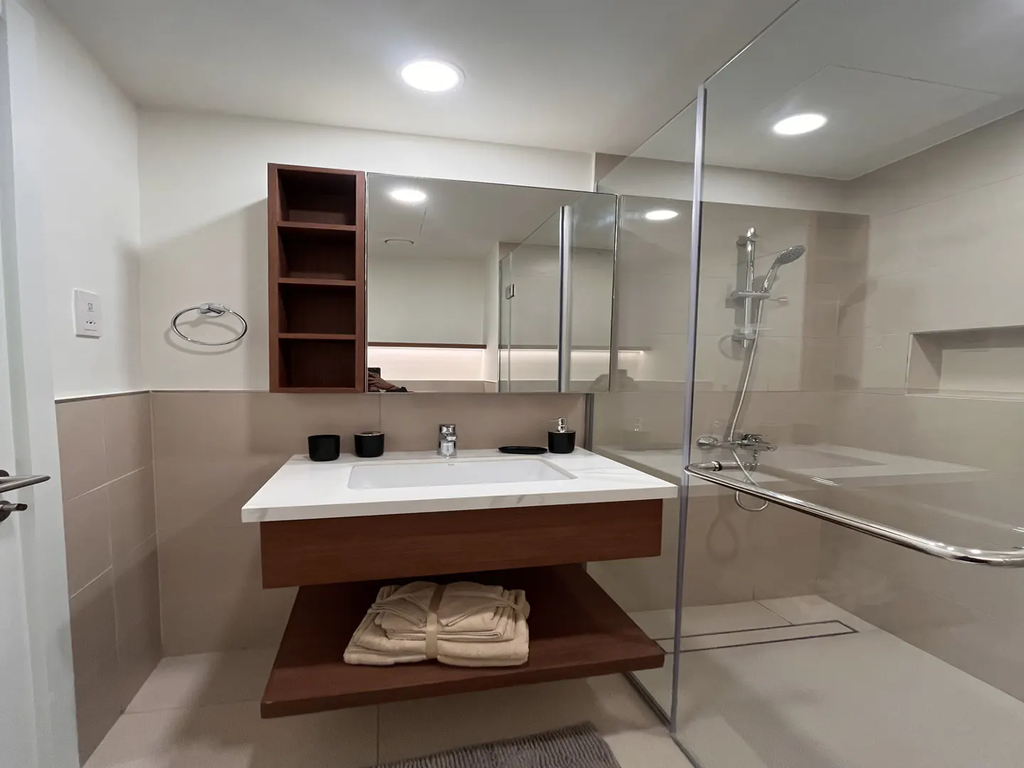 Modern bathroom with a white sink, wood vanity, and glass-enclosed shower. Towels are neatly stacked on a shelf.