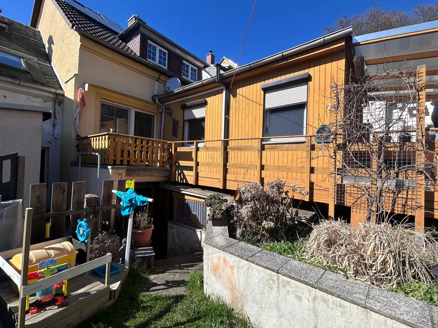 Backyard view of a yellow house with a wooden deck and a small garden with children's toys. Blue sky.