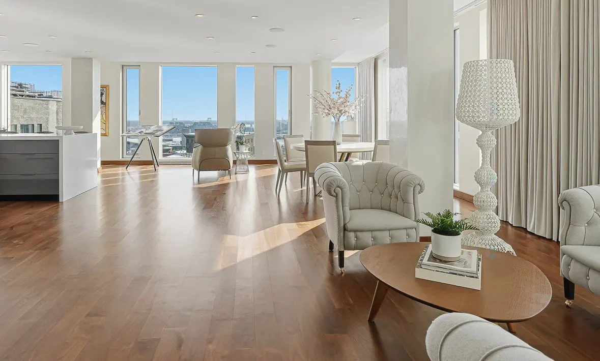 Bright, open living space with hardwood floors, large windows, and city views. Features tufted armchairs, a dining table, and a modern kitchen island.