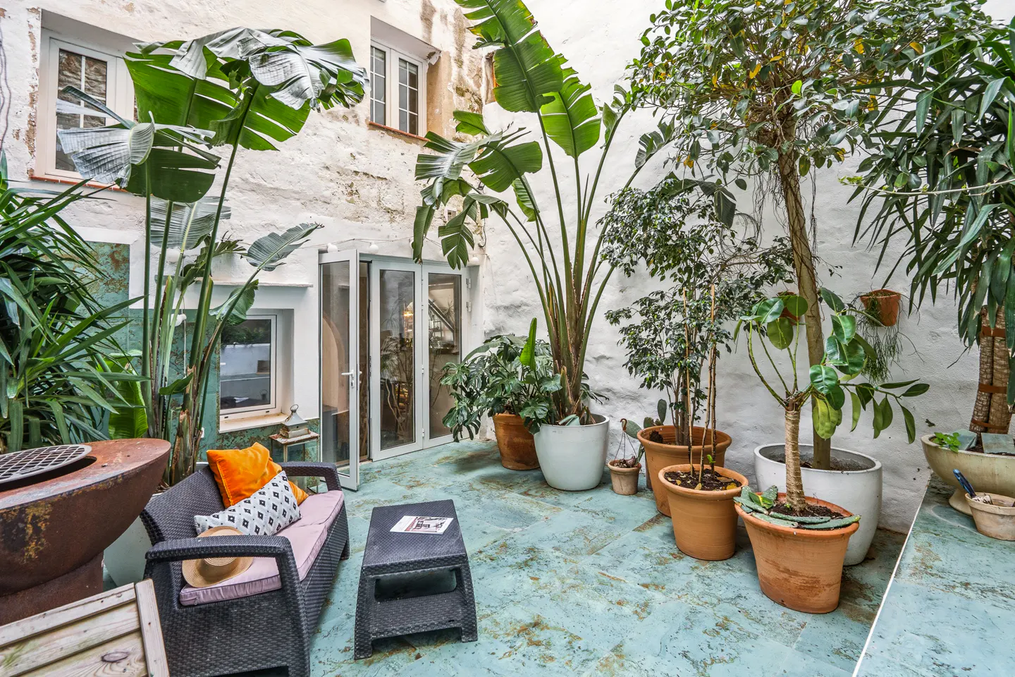 Outdoor patio with plants, sofa, and table. White walls and blue floor. The sofa has orange and patterned pillows. A hat sits on the sofa.