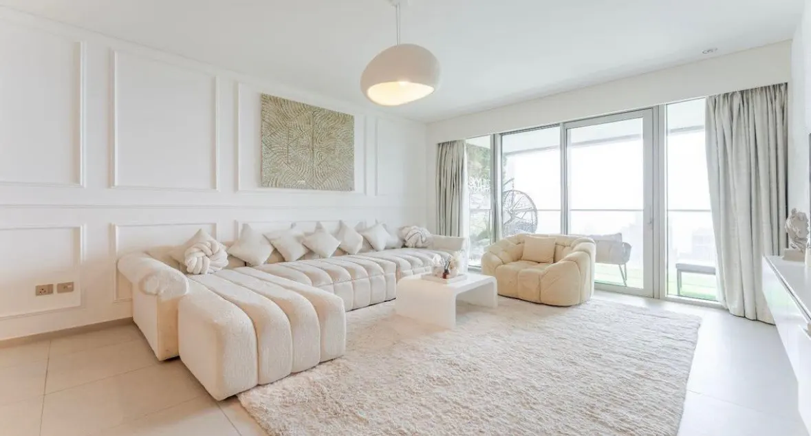 Bright, all-white living room with a large sectional sofa, fluffy rug, and a view of the balcony through sliding glass doors.