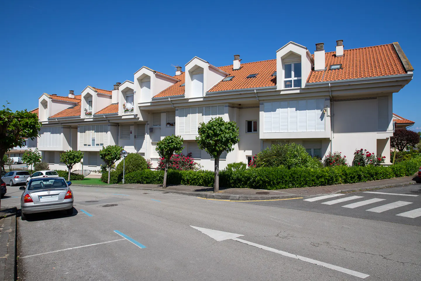 Street view of modern, white townhouses with orange tile roofs under a clear blue sky. Cars parked on the street.