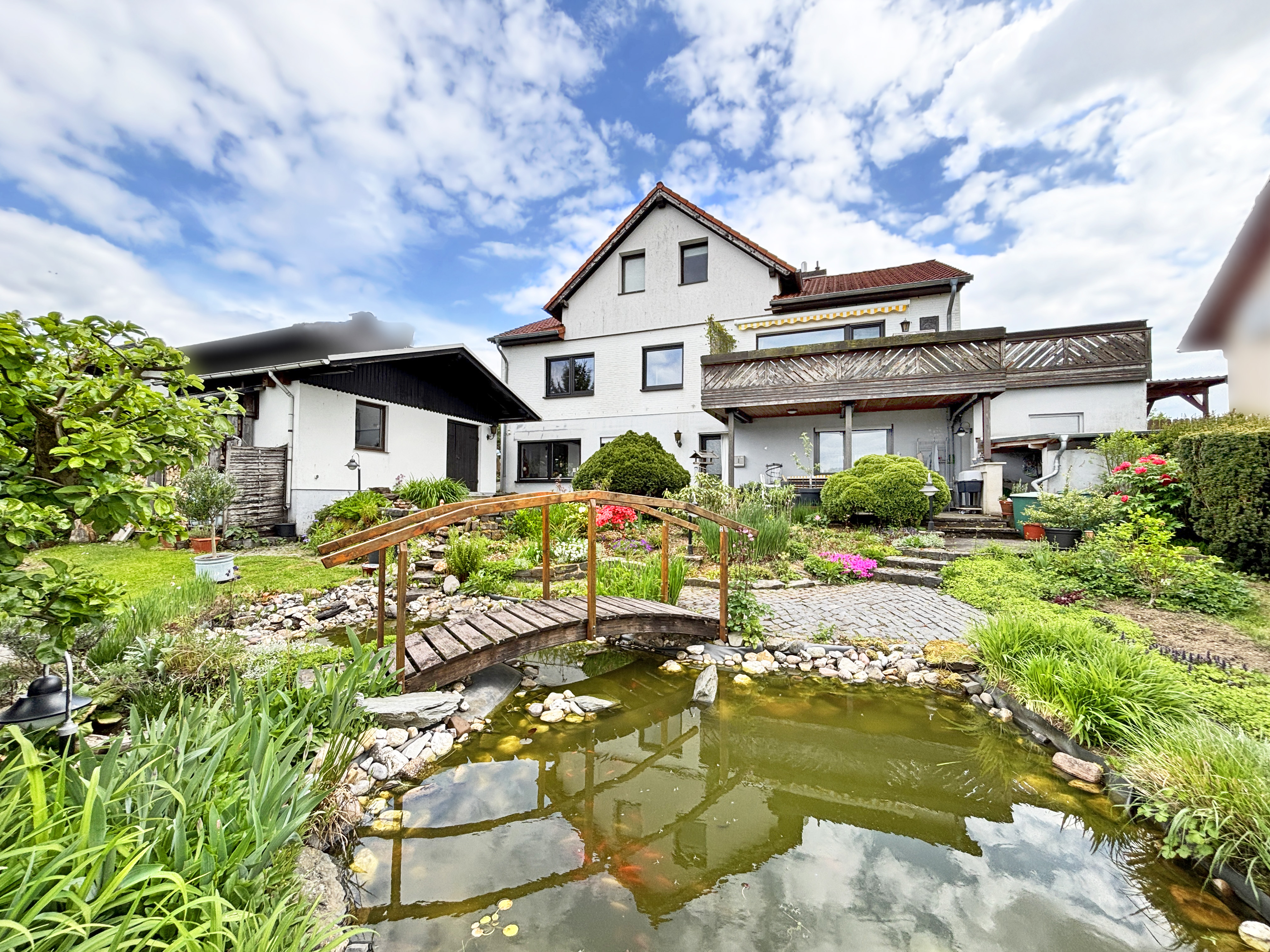 A white house with a red roof overlooks a garden with a pond and wooden bridge. The sky is blue with white clouds.