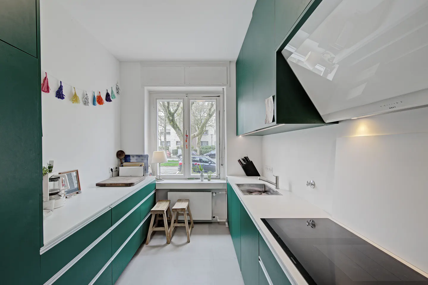 A modern, narrow kitchen with green cabinets, white countertops, and a window view. Two wooden stools sit near the window. A colorful tassel garland hangs on the wall.
