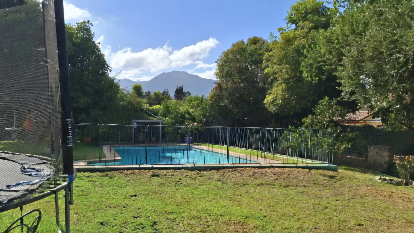 Backyard view with a blue pool surrounded by a fence, green grass, trees, and a mountain in the background under a blue sky with white clouds.