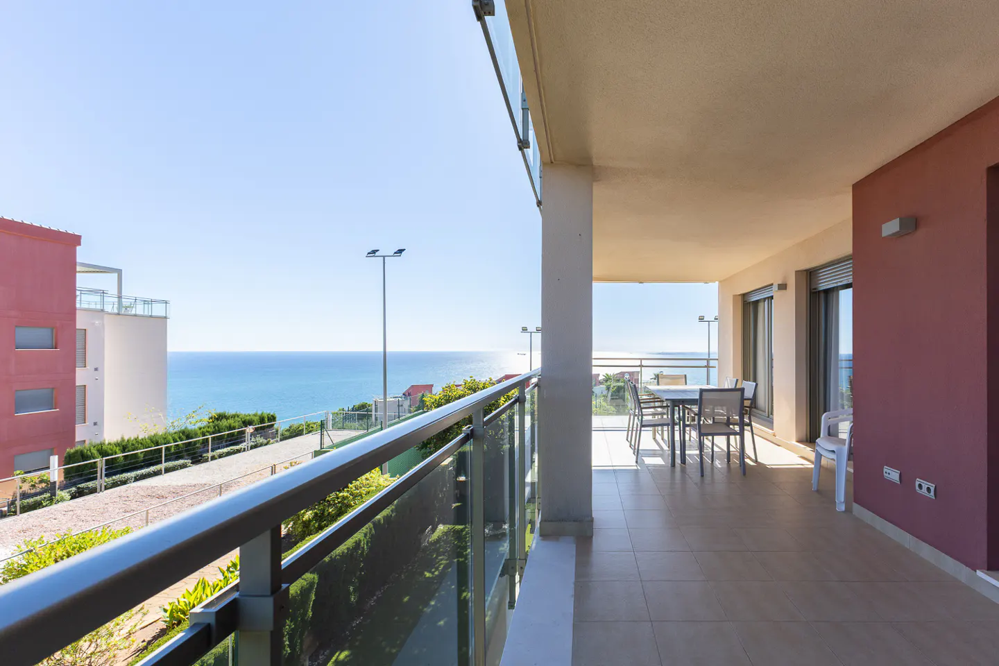 Balcony view of the ocean. Table and chairs on the patio. Glass railing. Blue sky.