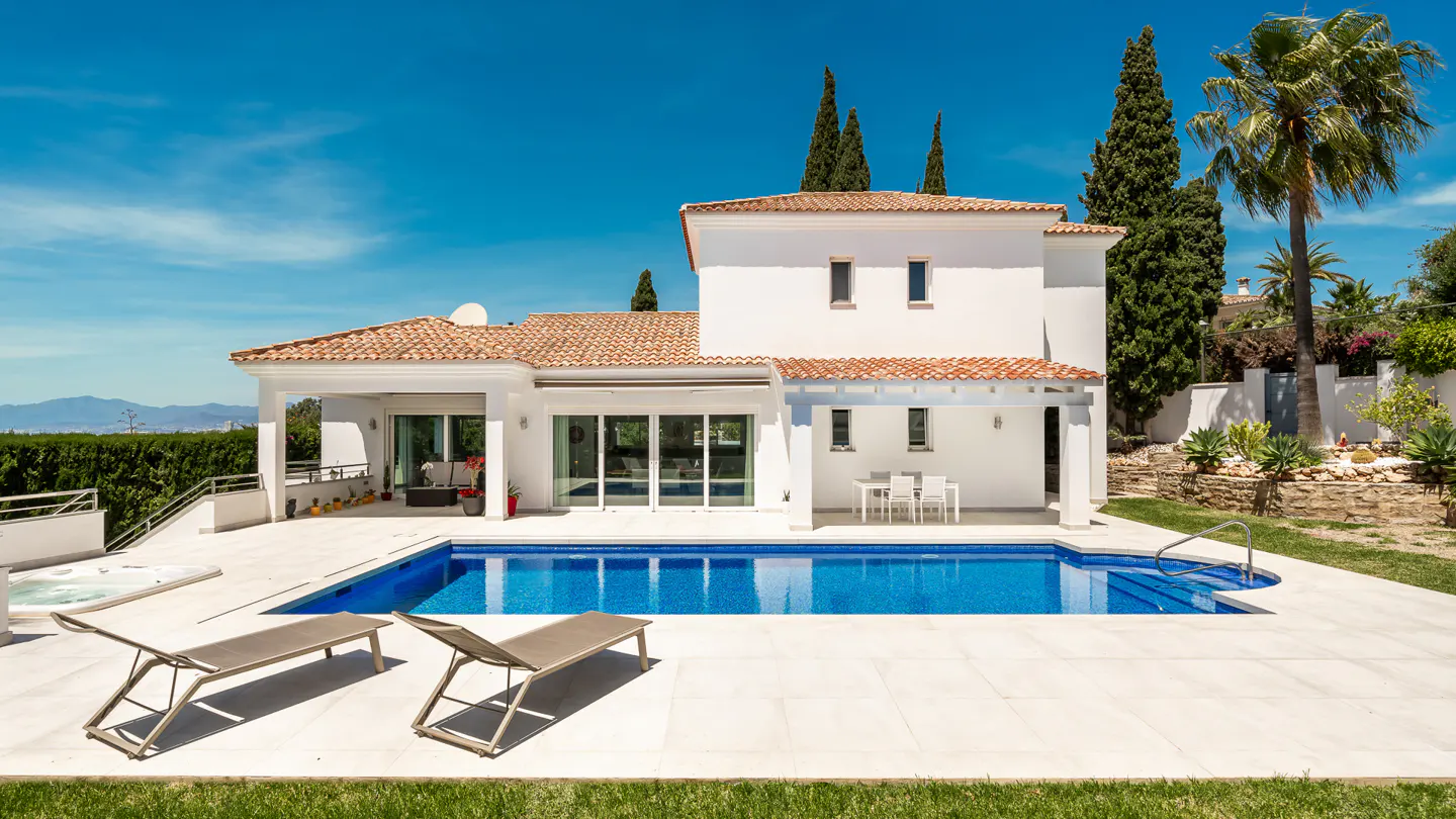 A white two-story house with a red tile roof, a blue pool, and two lounge chairs.